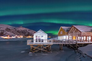 A wooden structure with a glass enclosure, a wooden building with a sloped roof, and a snow-covered landscape under a night sky with aurora borealis. - Sommarøy Arctic Hotel Tromsø (Tromsø)