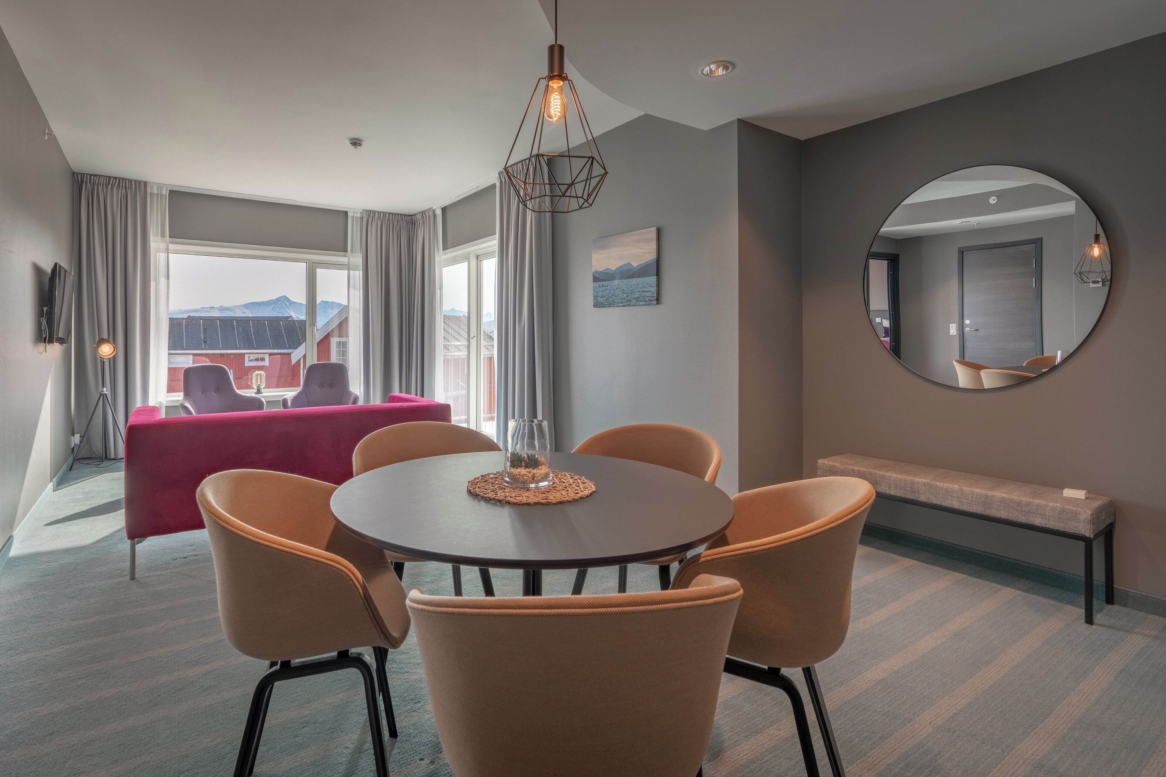 A modern hotel room with a round dining table, orange chairs, a red sofa, a bench, and a large mirror on the wall.