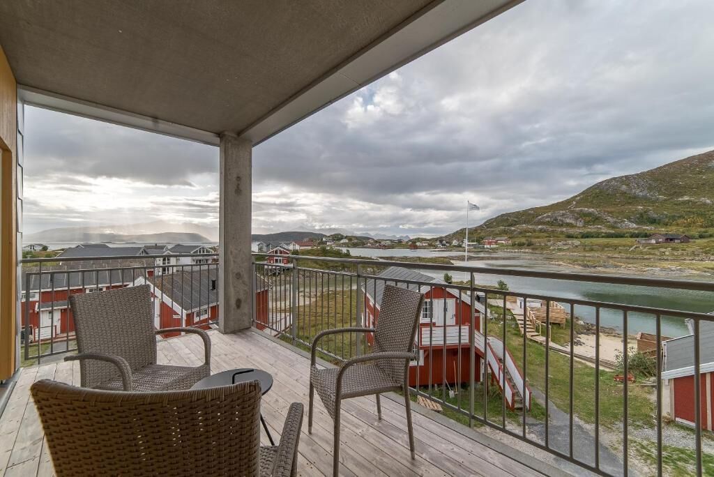 Un balcón con sillas de mimbre y una mesa, con vistas a un río y un edificio rojo con una escalera.