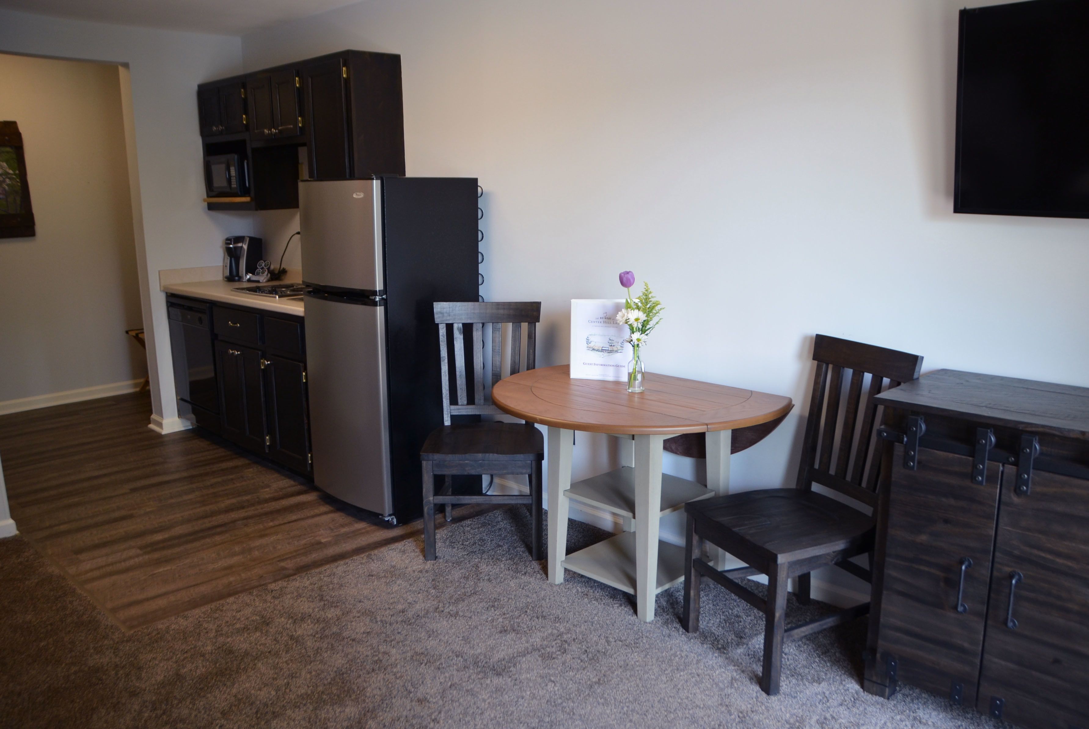 A compact kitchen with dark cabinets, a refrigerator, a microwave, and a coffee maker. There is a wooden table with two chairs and a vase with flowers.