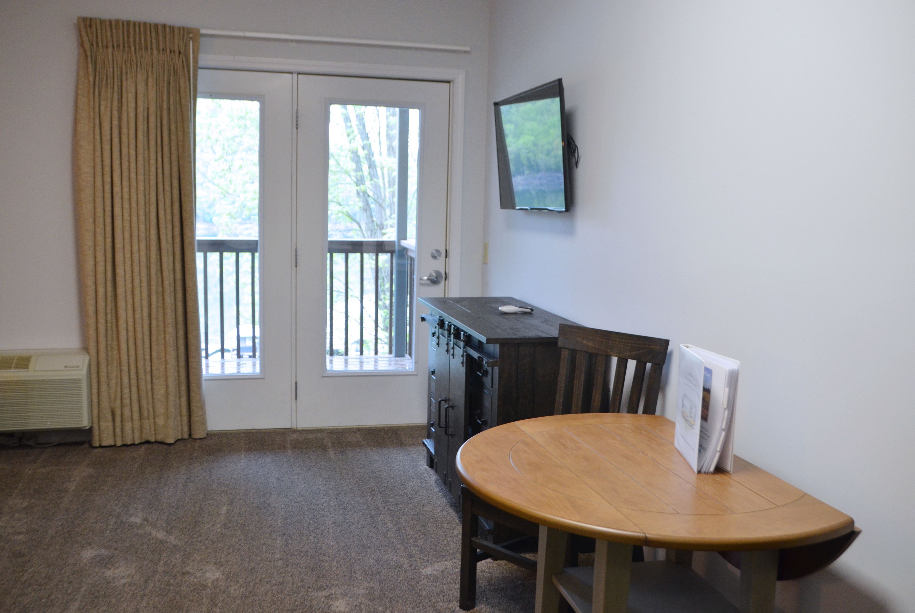 A room with a wooden dining table, a TV mounted on the wall, and a sliding glass door leading to an outdoor balcony.