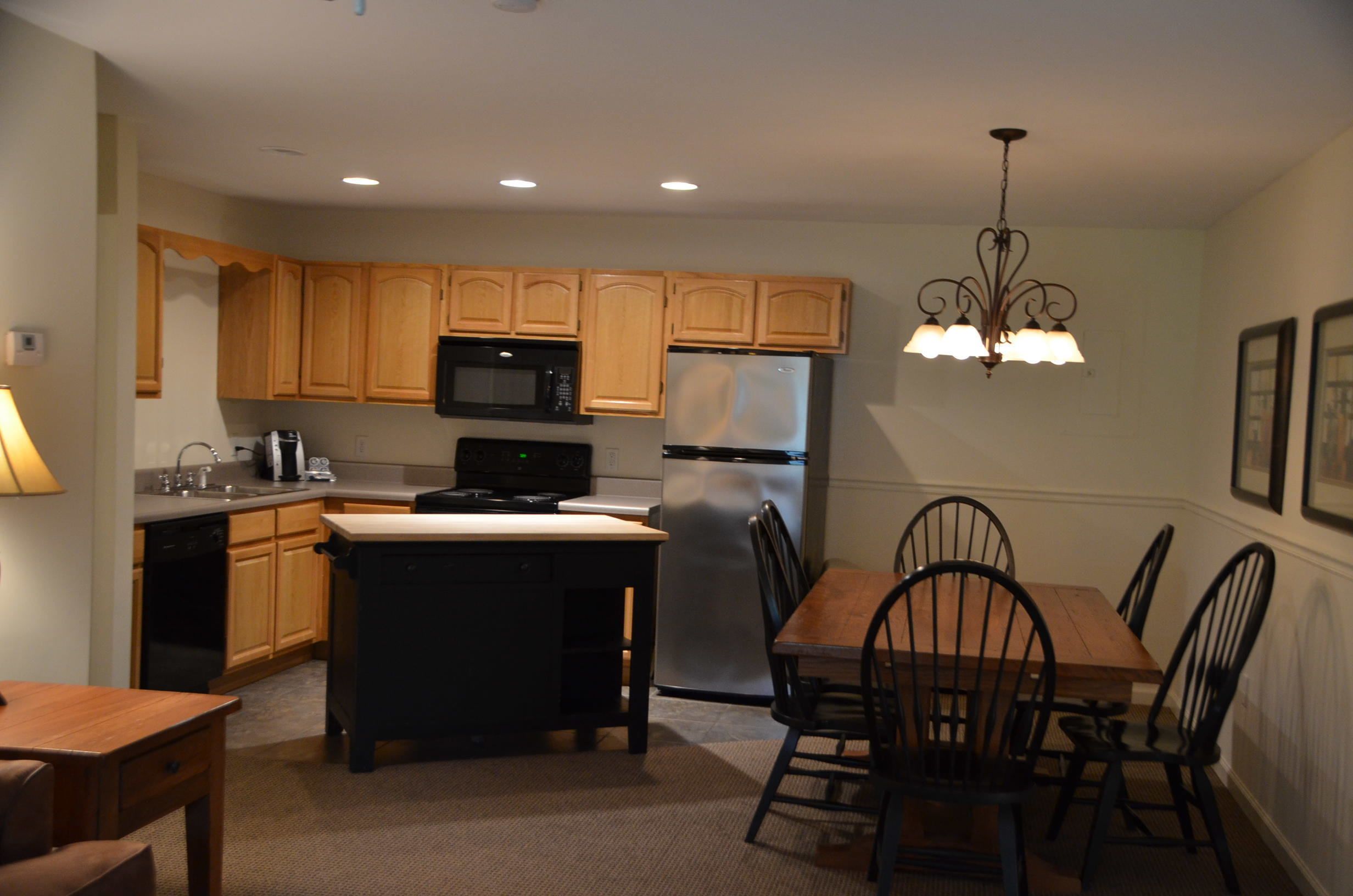 A compact kitchen with wooden cabinets, a microwave, a refrigerator, a dining table with chairs, and a chandelier.