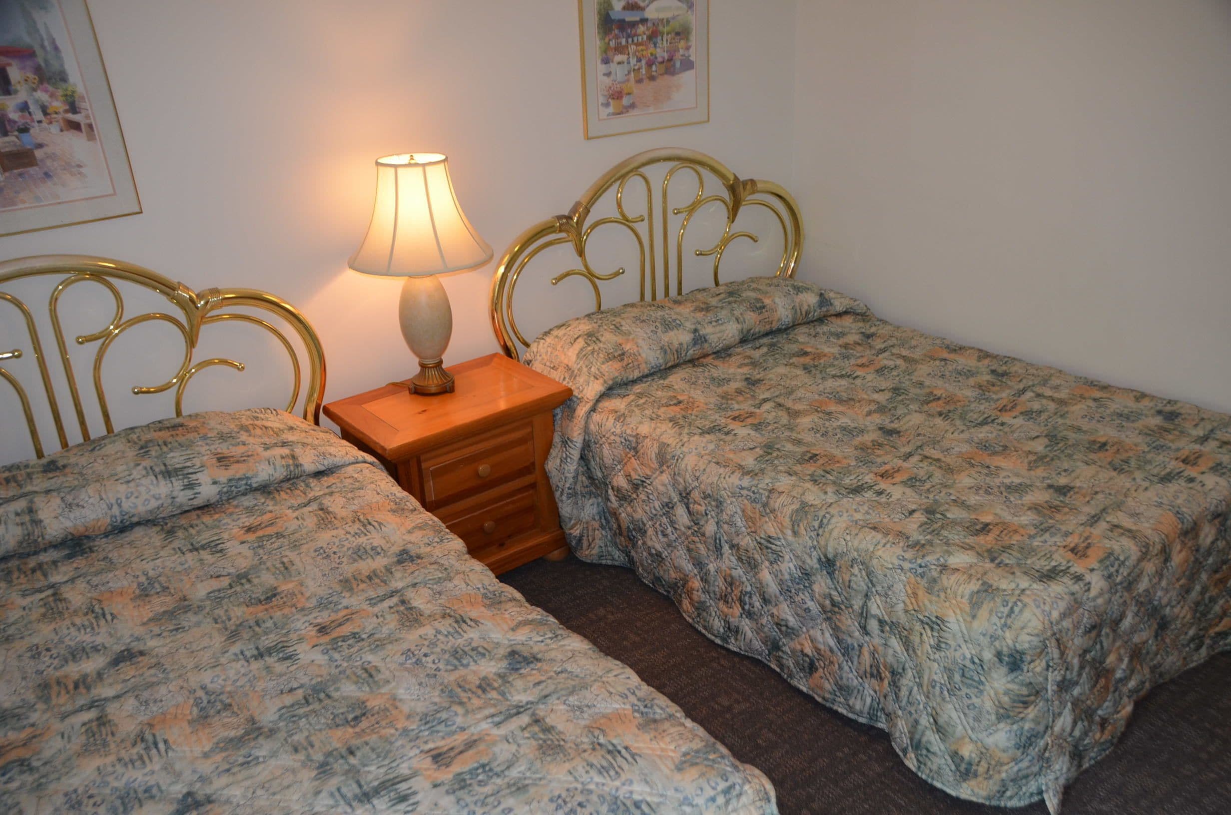 A room with two beds, a nightstand with a lamp, and framed pictures on the wall.