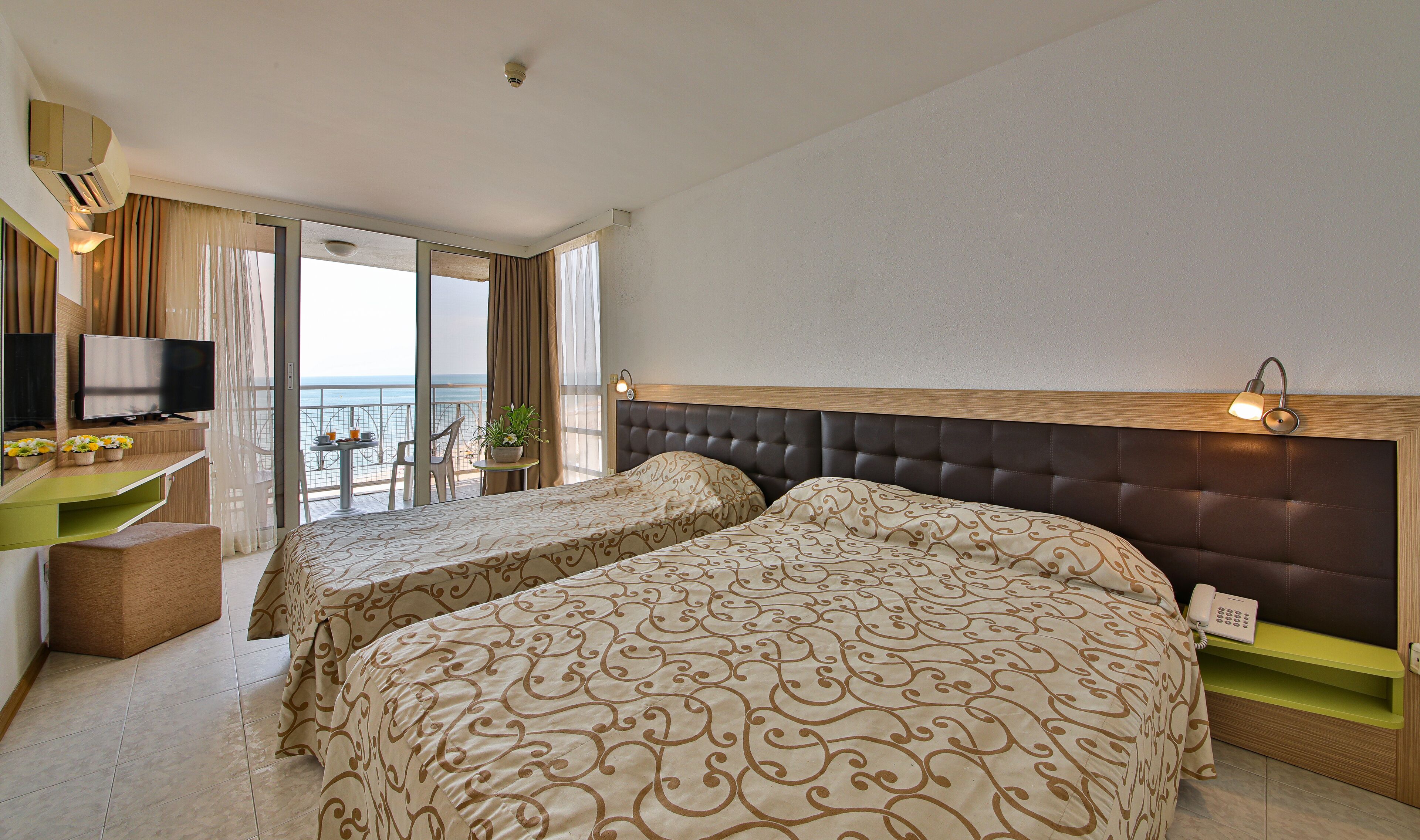 A hotel room with two beds, a TV, a small table with drinks, and a balcony with a view of the sea.