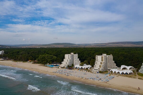 A beach resort with multiple white buildings, a sandy beach with numerous umbrellas, and a forested background.