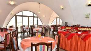 A restaurant with round tables covered in white tablecloths and orange tablecloths, wooden chairs, and a large window in the background.