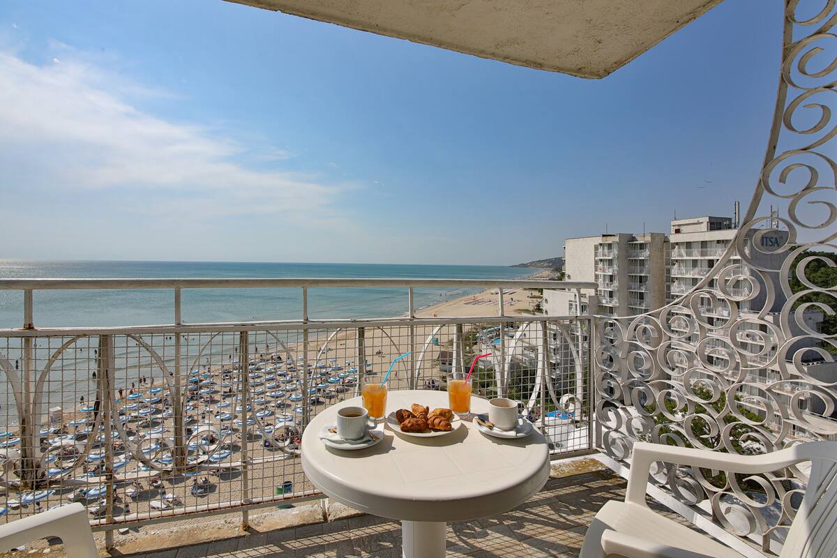 Varanda com mesa posta para o café da manhã, com vista para uma praia movimentada com guarda-sóis e pessoas.