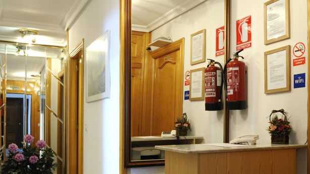 A reception area with a wooden reception desk, a fire extinguisher, framed certificates, and a small plant. - Iberos Jaén (Jaén)