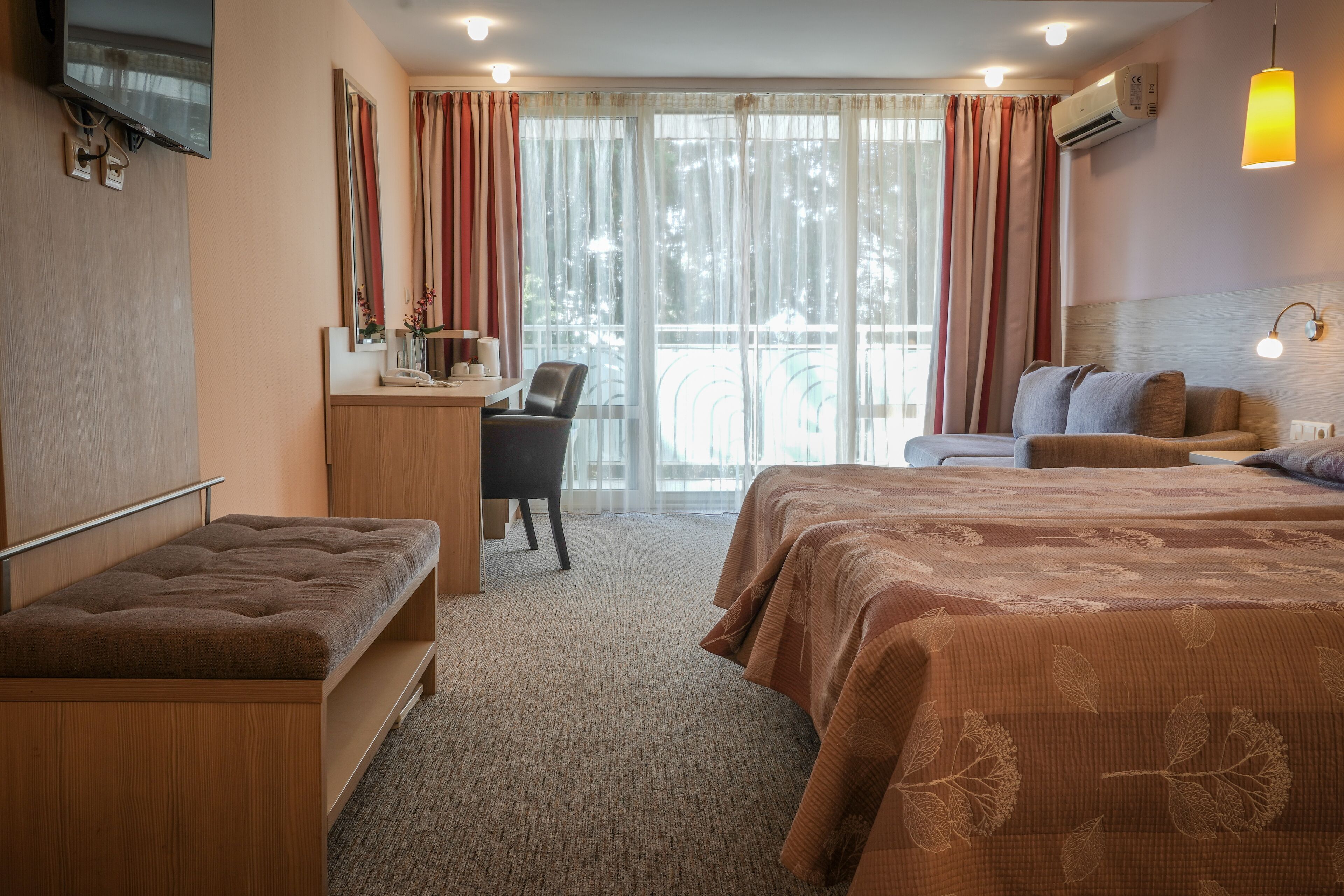 Quarto de hotel com cama, escrivaninha, cadeira, banco e uma grande janela com cortinas.