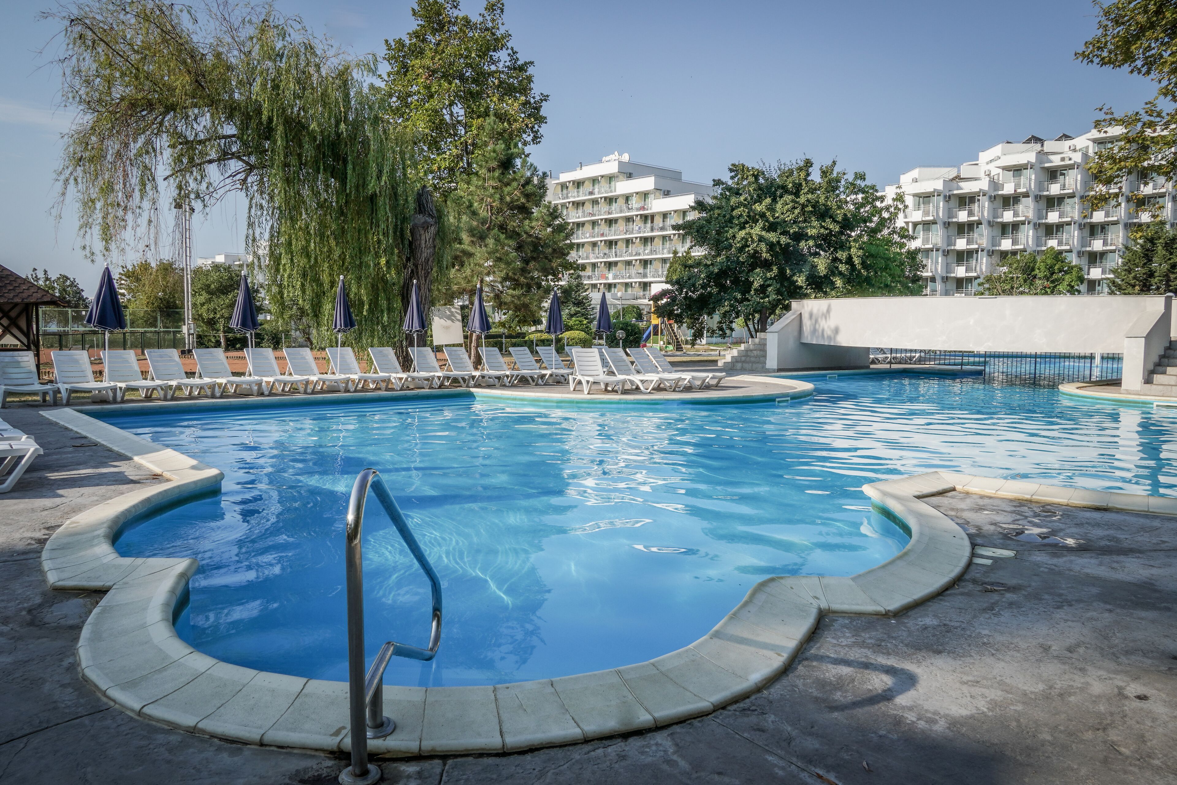 A swimming pool with lounge chairs and umbrellas, surrounded by trees and a building in the background.
