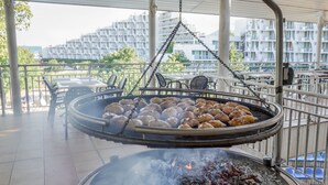 A barbecue grill with food being cooked, surrounded by outdoor seating and a view of a residential building in the background. - Hotel Sandy Beach (Albena)