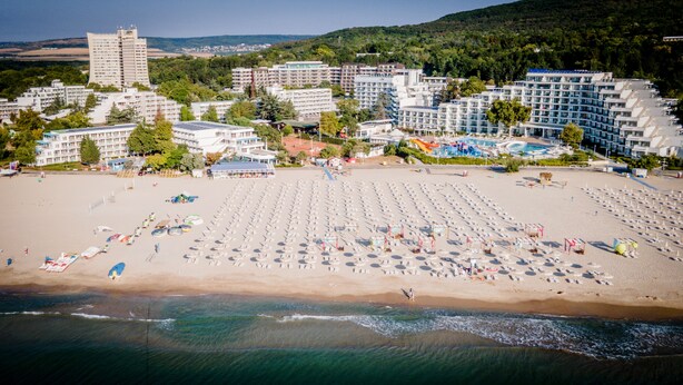 A beach with rows of sunbeds and umbrellas, a pool area, and buildings in the background. - Hotel Sandy Beach (Albena)