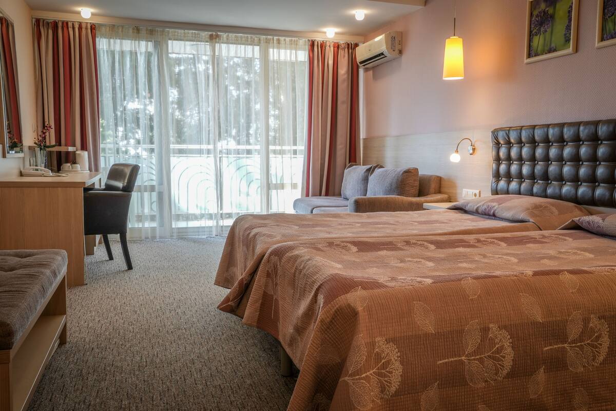 A hotel room with a large bed, a desk with a chair, a sofa, and a balcony with curtains.