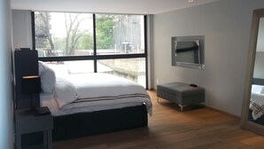 A modern bedroom with a large bed, a bedside table, a bench, a TV mounted on the wall, and a large window offering a view of the outdoors. - Polanco by O (Mexico City)