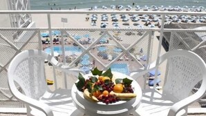 Un balcon avec des chaises blanches et une table sur laquelle se trouvent des fruits et une bouteille. La vue comprend une plage avec des parasols et des personnes.