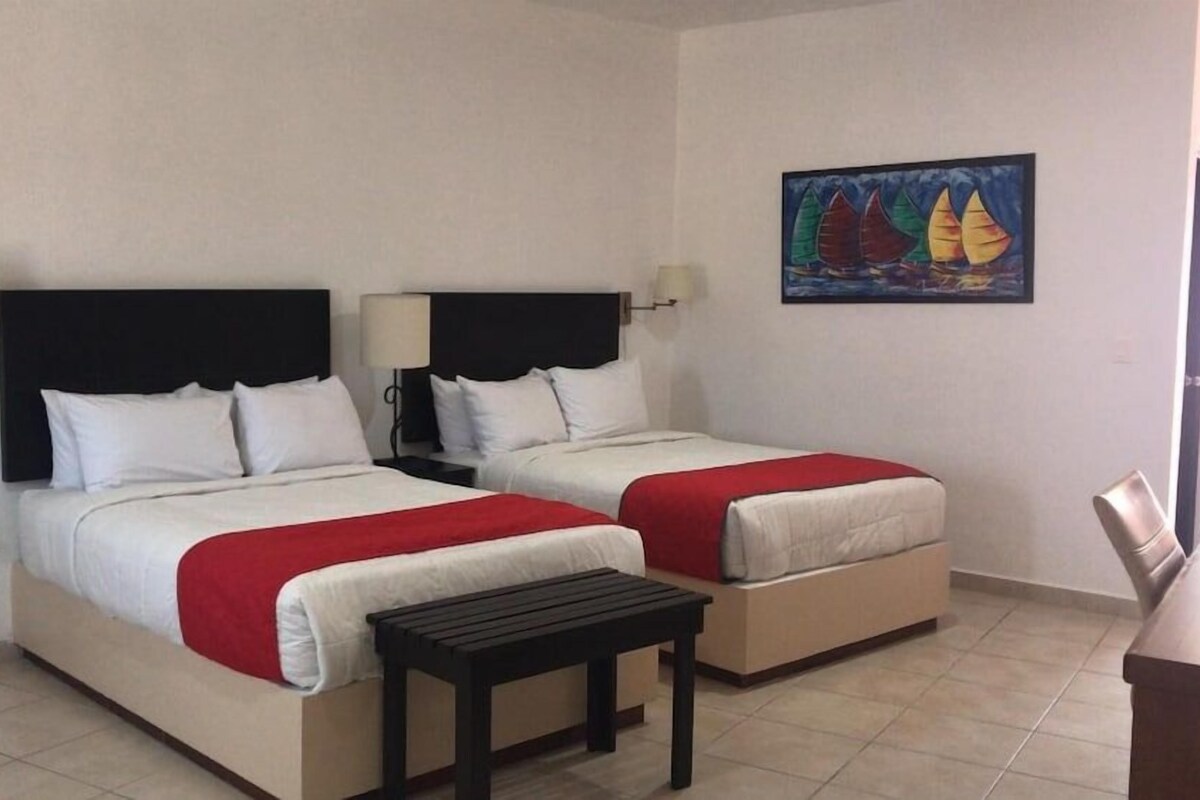 Habitación de hotel con dos camas, cada una con cabecero negro, ropa de cama blanca y manta roja. Hay una mesita de noche frente a las camas y un cuadro en la pared.