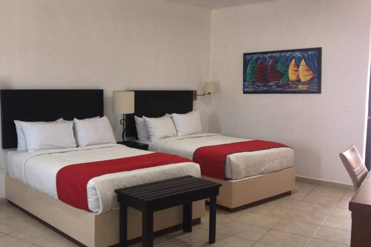 A room with two beds, each with white bedding and red accents, a small table, and a painting on the wall.