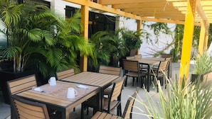 An outdoor patio area with wooden tables and chairs, surrounded by potted plants and a yellow pergola structure. - Hotel Marina Topolobampo (Topolobampo)