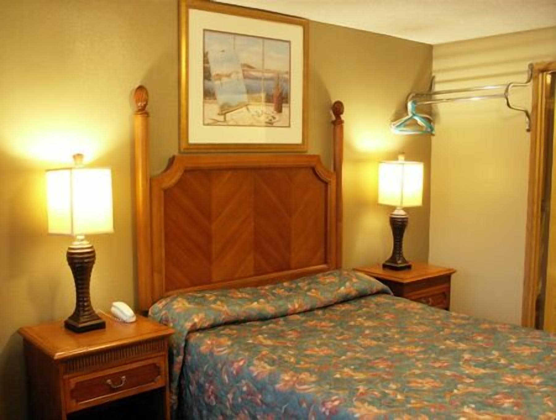 Een hotelkamer met een houten hoofdbord, twee bedlampen, een bloemenbedekking, een ingelijst schilderij aan de muur en een raam met gordijn.
