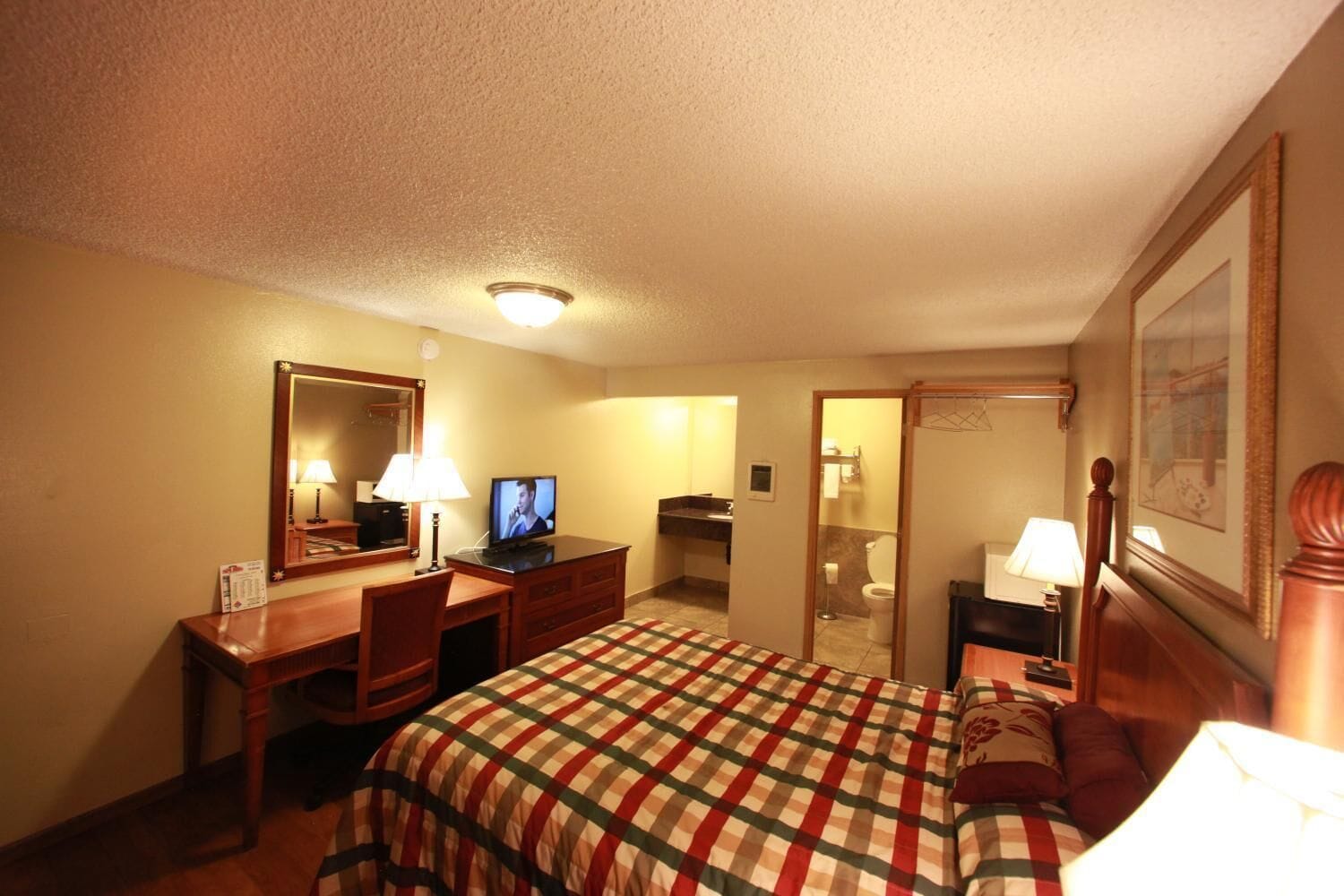 Habitación de hotel con una cama, un escritorio con computadora, televisión, espejo y baño visible a través de una puerta abierta.