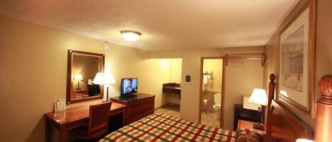 Habitación de hotel con una cama, un escritorio con computadora, televisión, espejo y baño visible a través de una puerta abierta.