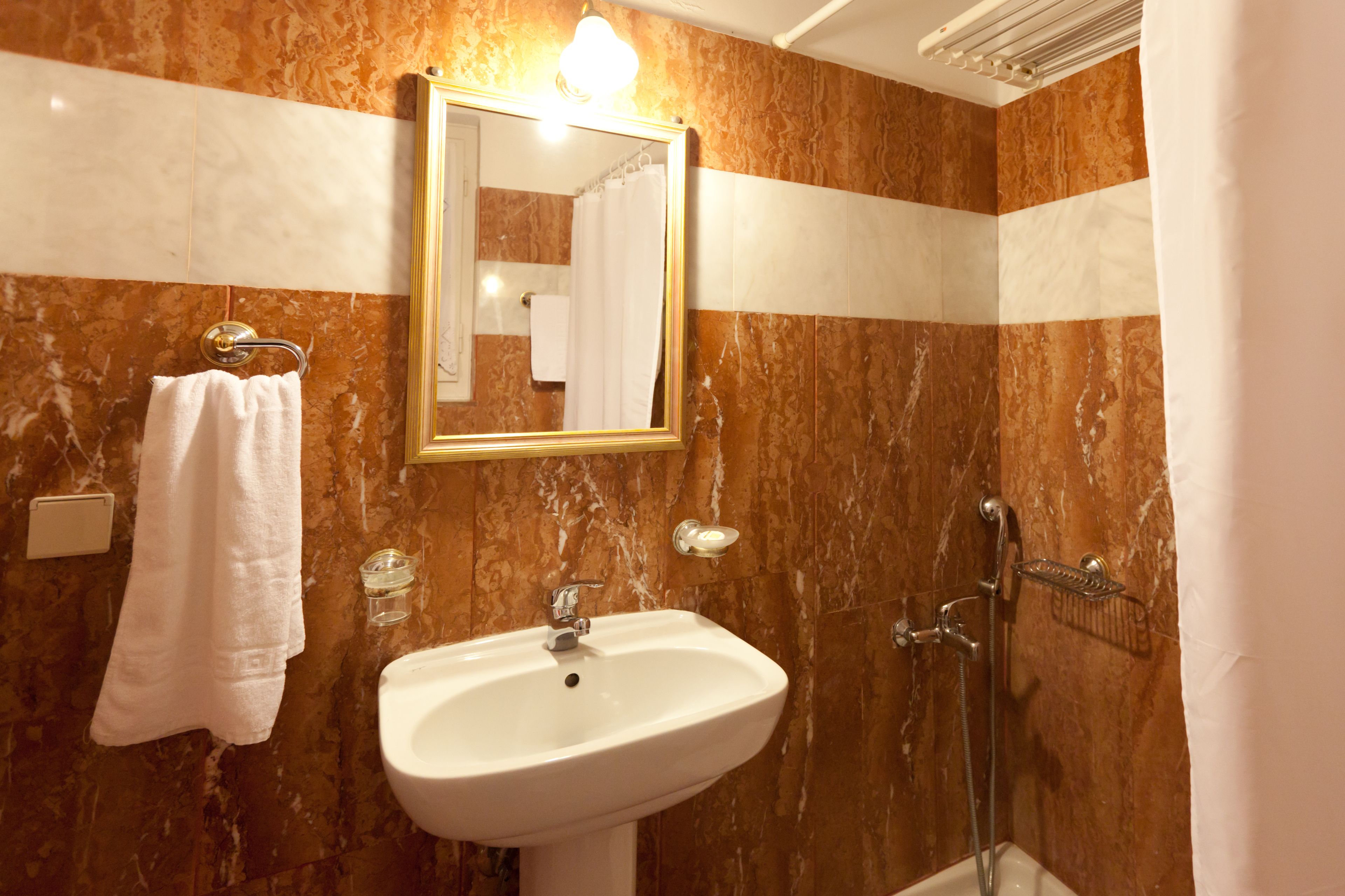 A bathroom with a marble wall, a sink, a shower, and a mirror with a gold frame.