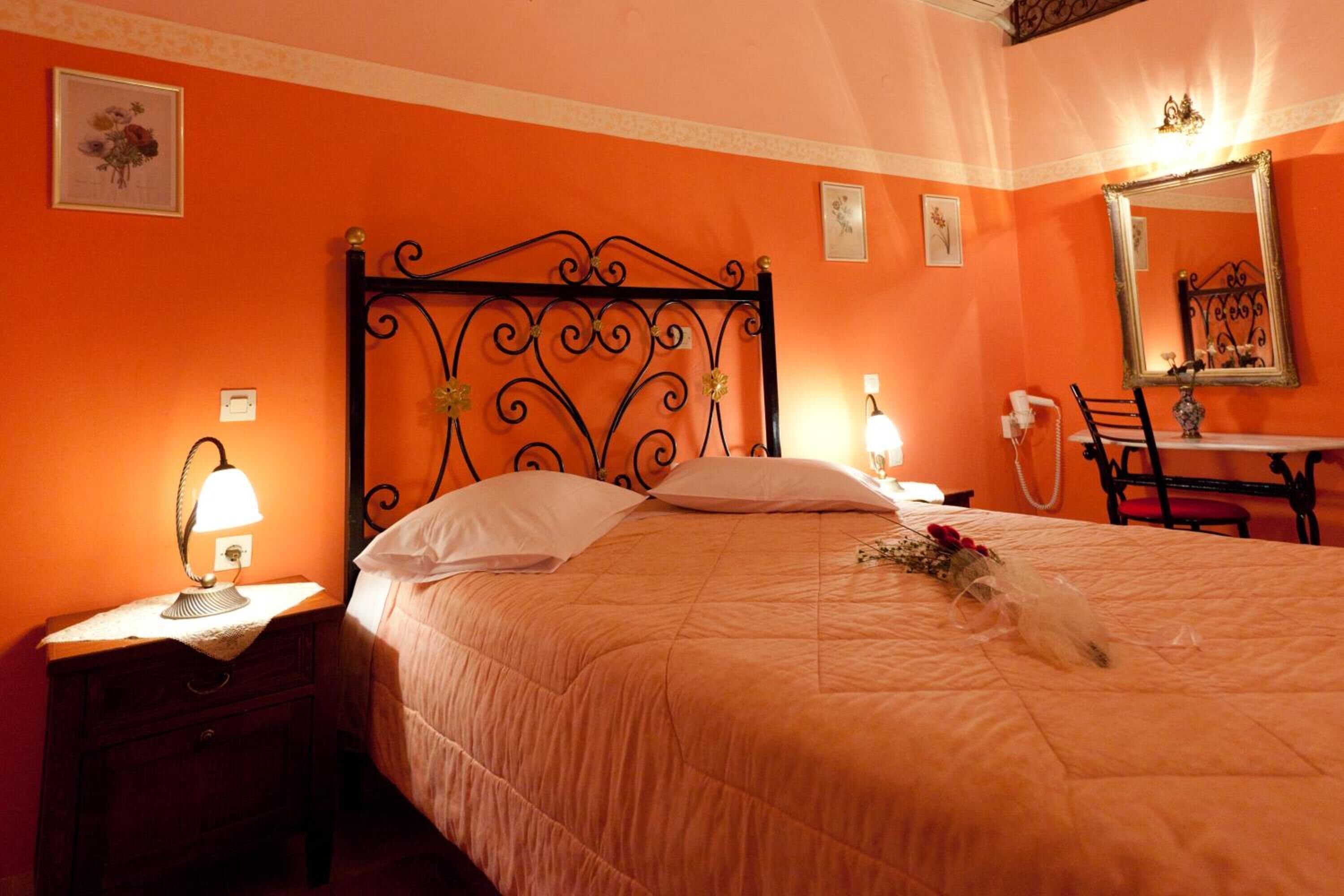 A hotel room with a bed, a nightstand with a lamp, a desk with a chair, and a mirror on the wall.