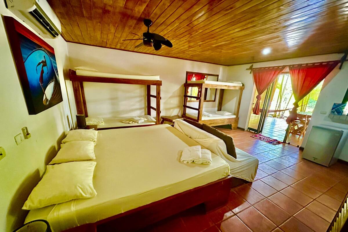 A room with a large bed, a bunk bed, a ceiling fan, and a small kitchenette.
