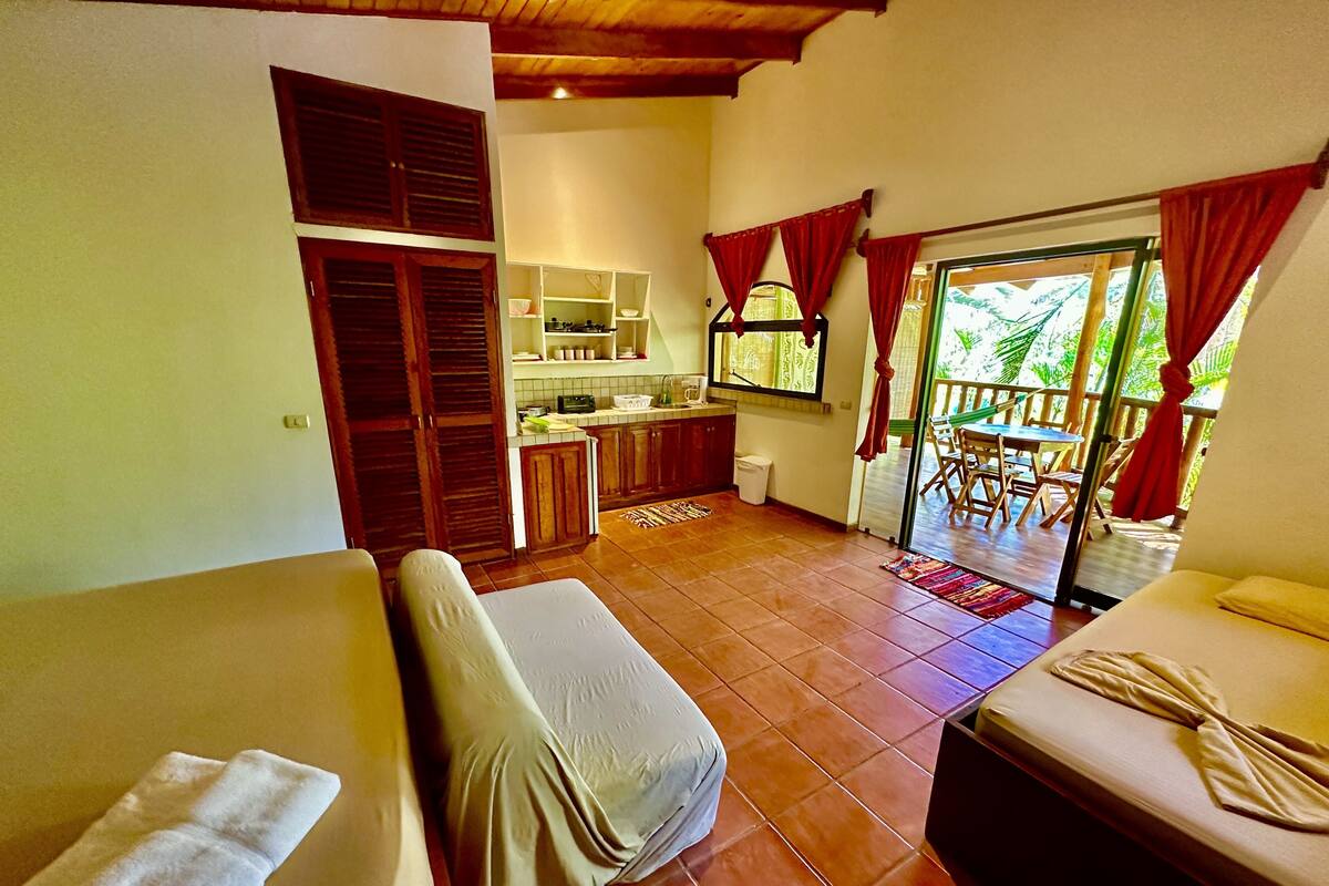 A room with a bed, a sofa, a small table, a kitchen area, and a balcony with red curtains.