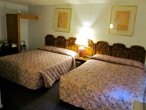 A hotel room with two double beds, a wooden headboard, a bedside table with a lamp, and two framed pictures on the wall.