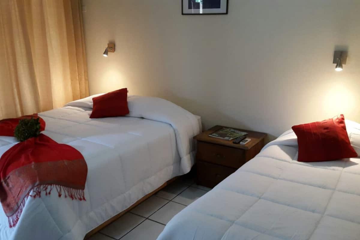 A hotel room with two beds, each with a red pillow and a blanket. There is a nightstand between the beds with a book and a lamp.