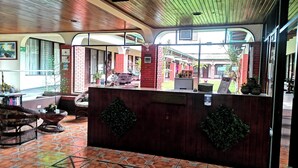 A reception area with a tiled floor, wooden reception desk, and a view of the outdoor area. - Hotel Wagelia Turrialba (Turrialba)