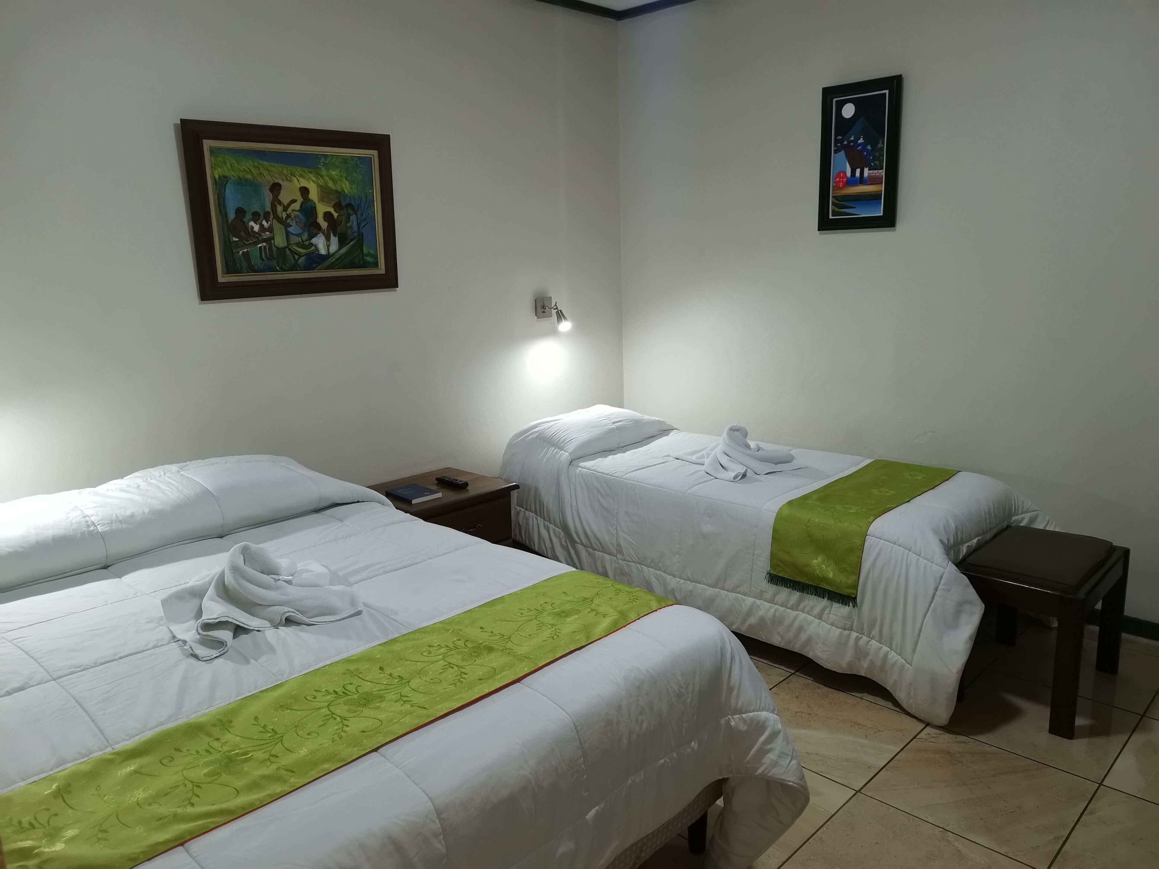 A hotel room with two beds, a nightstand, a painting, and a poster on the wall.