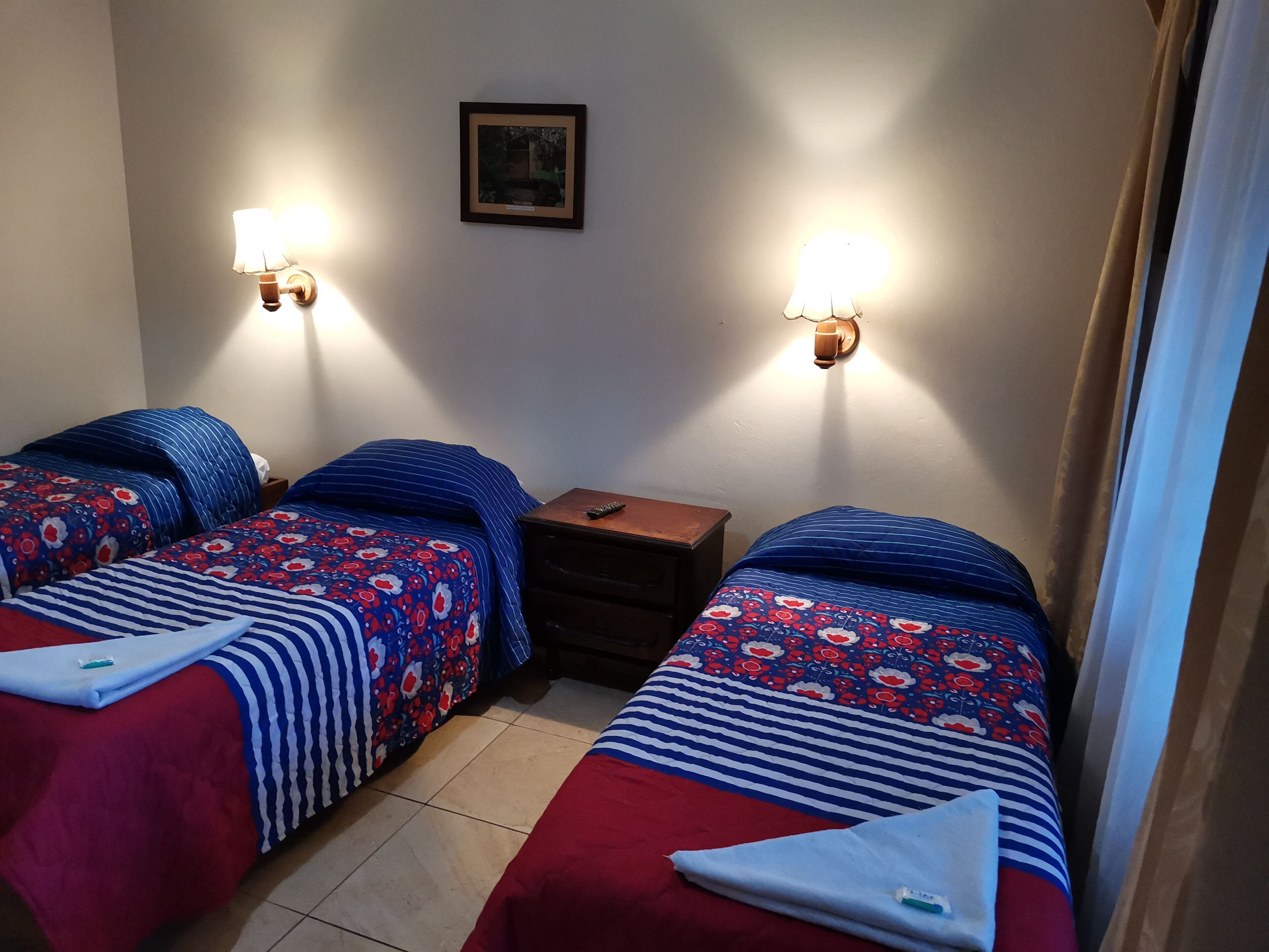 Una habitación con dos camas, cada una con una colcha de estampado floral y almohadas a rayas. Hay lámparas fijadas a la pared a cada lado de un cuadro enmarcado. Una mesita de noche de madera se encuentra entre las camas.