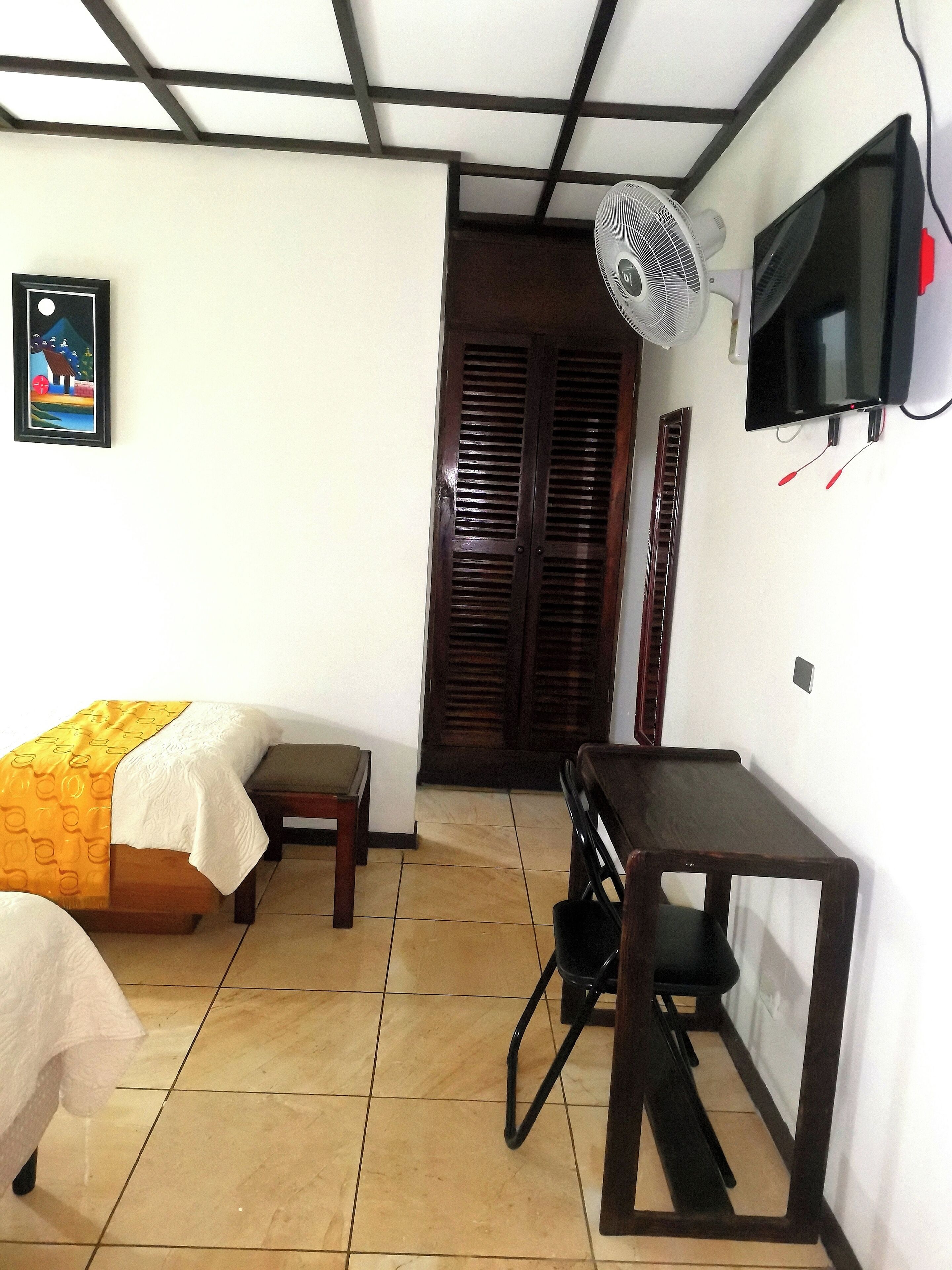 Quarto com cama, escrivaninha, cadeira, televisão e ventoinha.