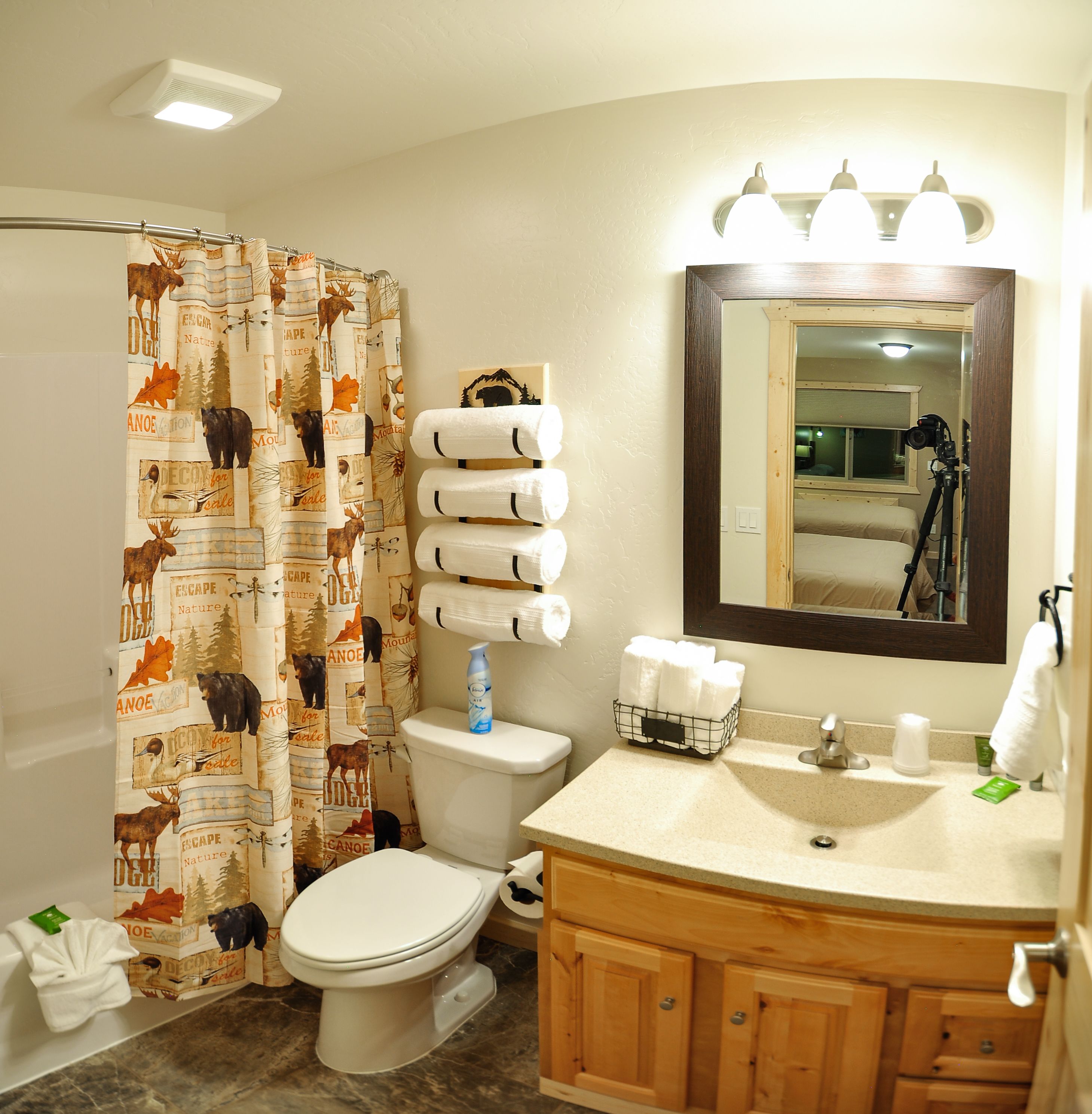 A bathroom with a toilet, sink, and mirror. There is a shower curtain with a nature print, a bottle of shampoo on the toilet, and a camera on the sink.