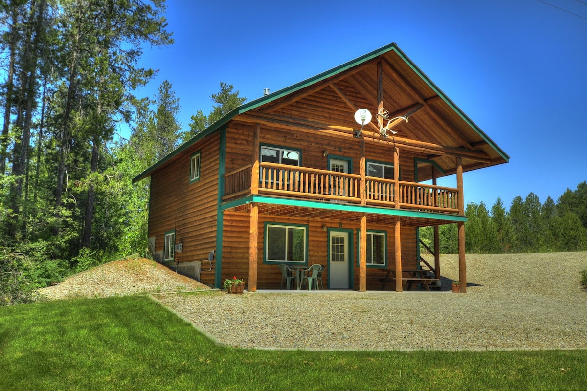 A two-story log cabin with a balcony, surrounded by a gravel driveway and lush greenery.