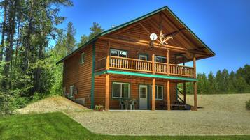 A two-story log cabin with a balcony, surrounded by a gravel driveway and lush greenery.