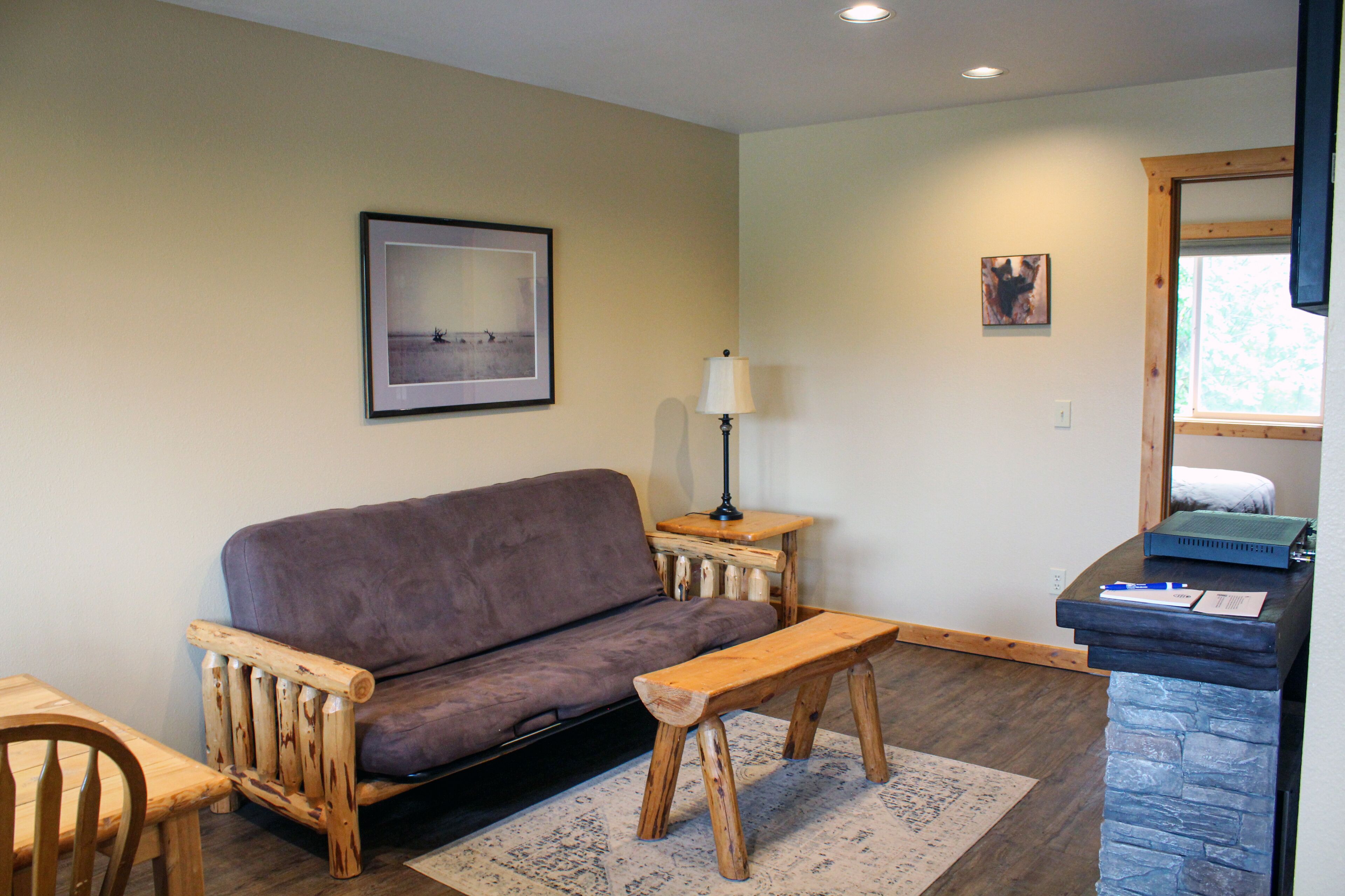 A cozy living room with a wooden sofa, a wooden bench, a small table, and a lamp. There is a framed picture on the wall and a window with a view of trees.