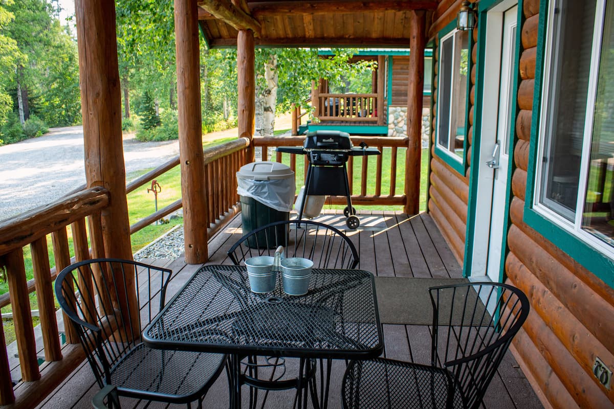 A covered outdoor area with a table and chairs, a grill, and a view of the surrounding forest.
