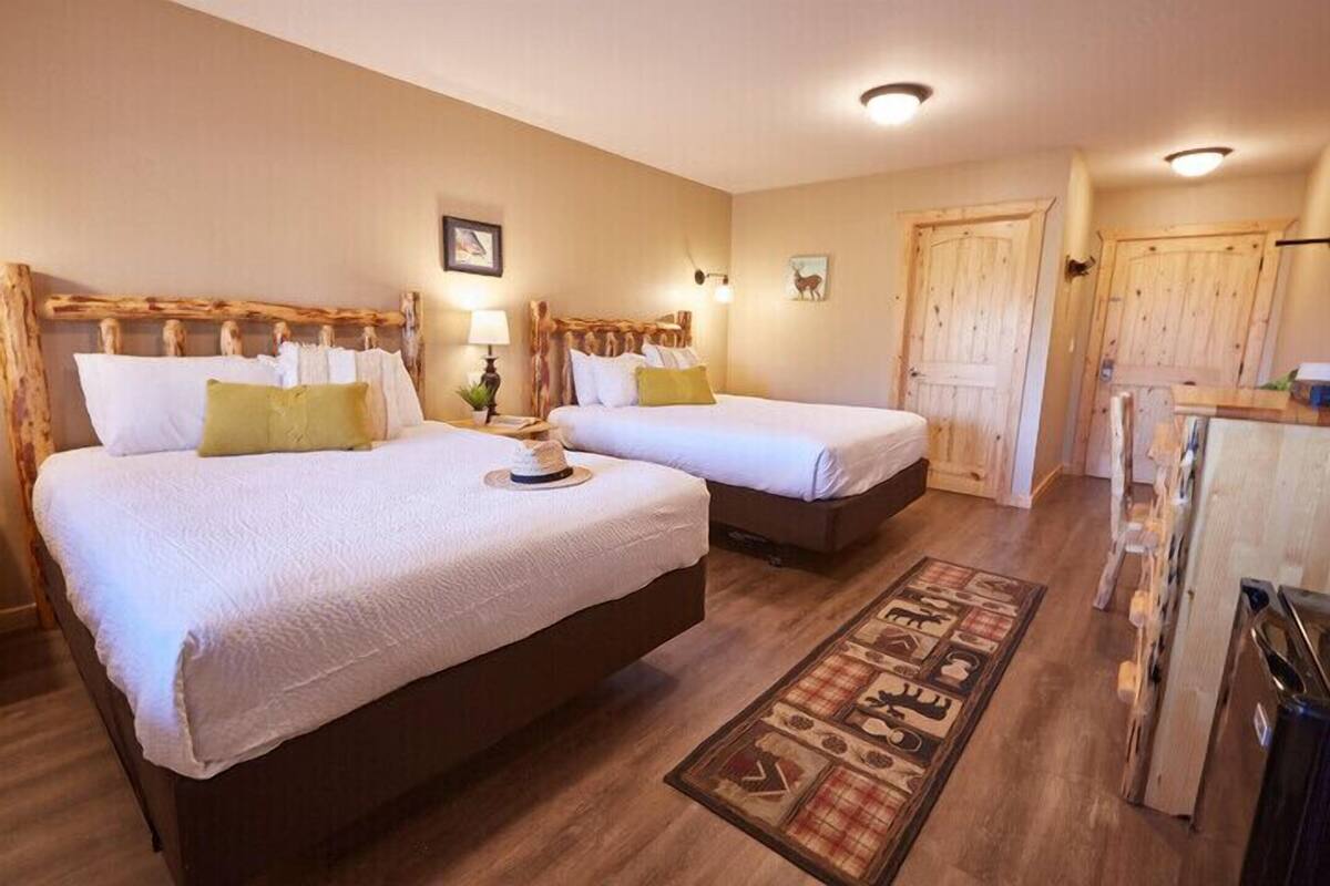 A hotel room with two beds, wooden headboards, a small table with a lamp, a wooden floor, and a door.