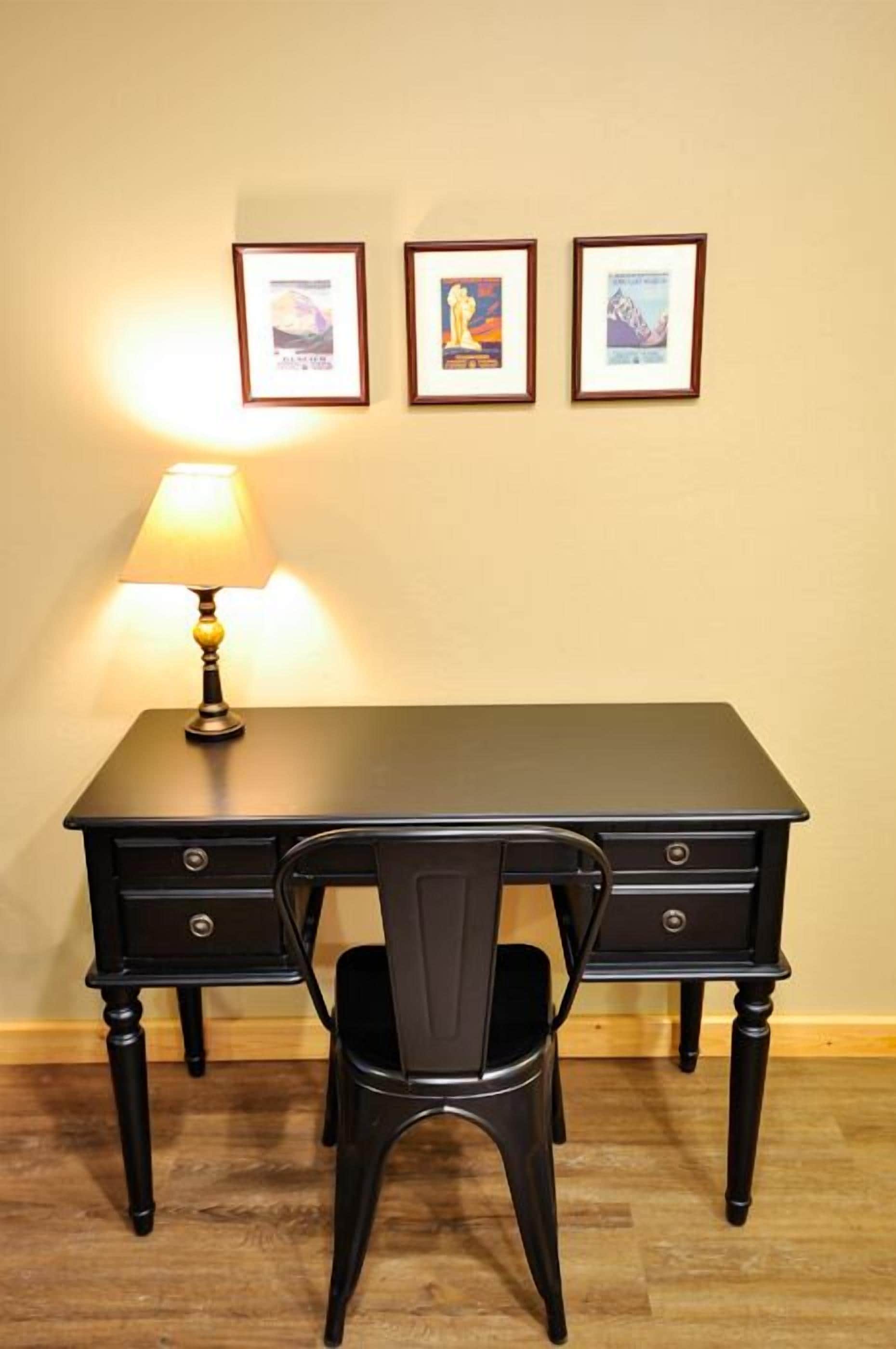 A dark wooden desk with a lamp, a chair, and three framed pictures on the wall.