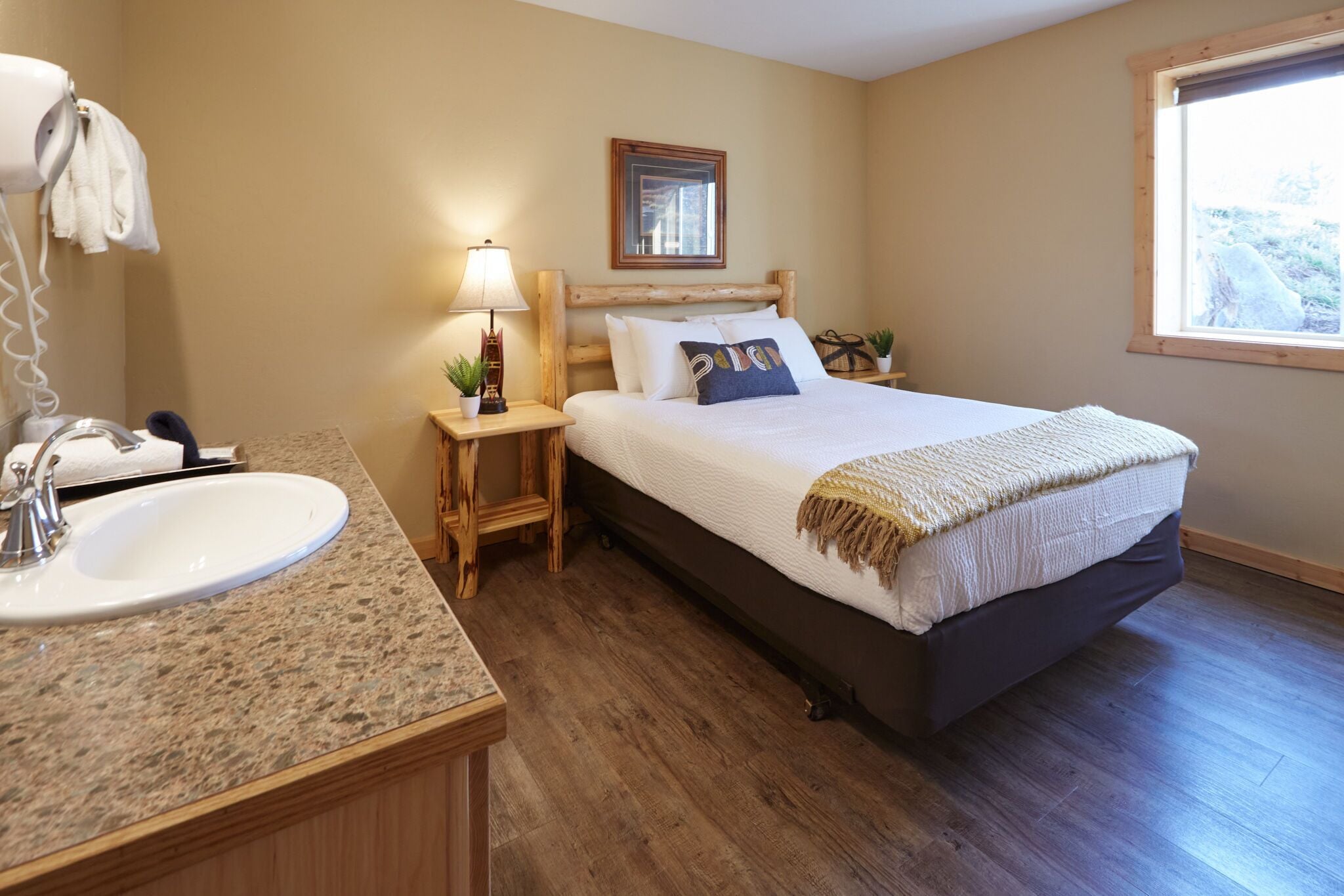 A bedroom with a bed, a bedside table with a lamp, a wooden headboard, a window, and a bathroom with a sink and mirror.
