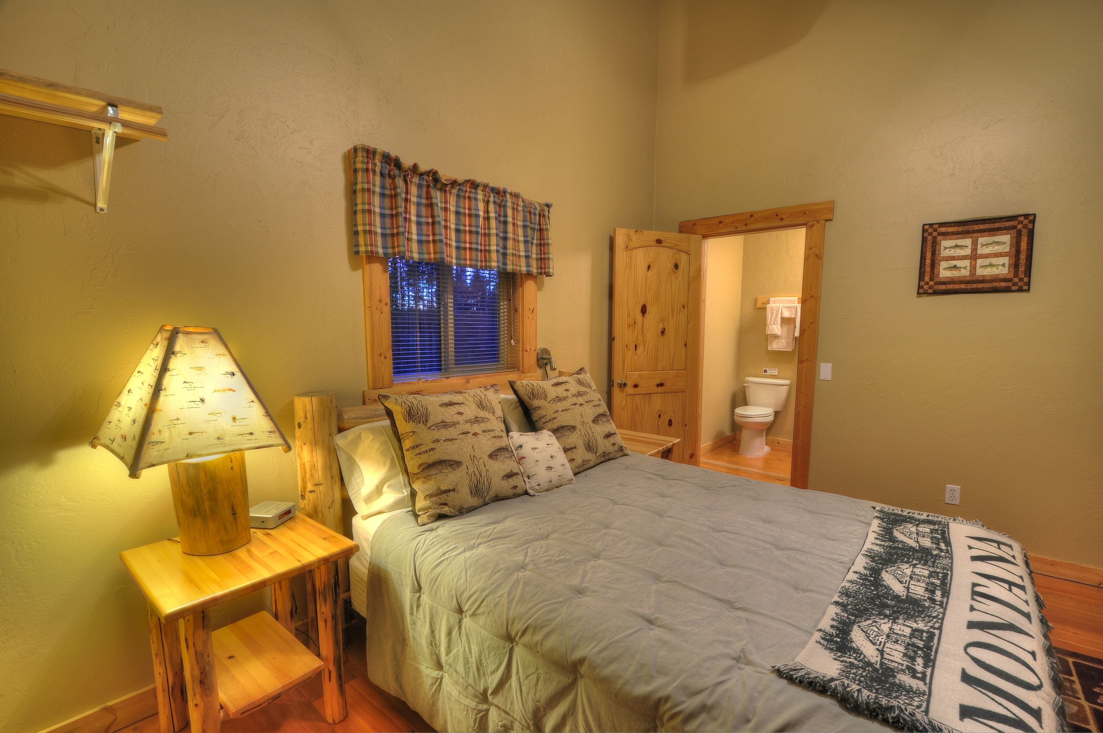 A bedroom with a bed, a nightstand with a lamp, a window with blinds, a wooden door, and a framed picture on the wall.
