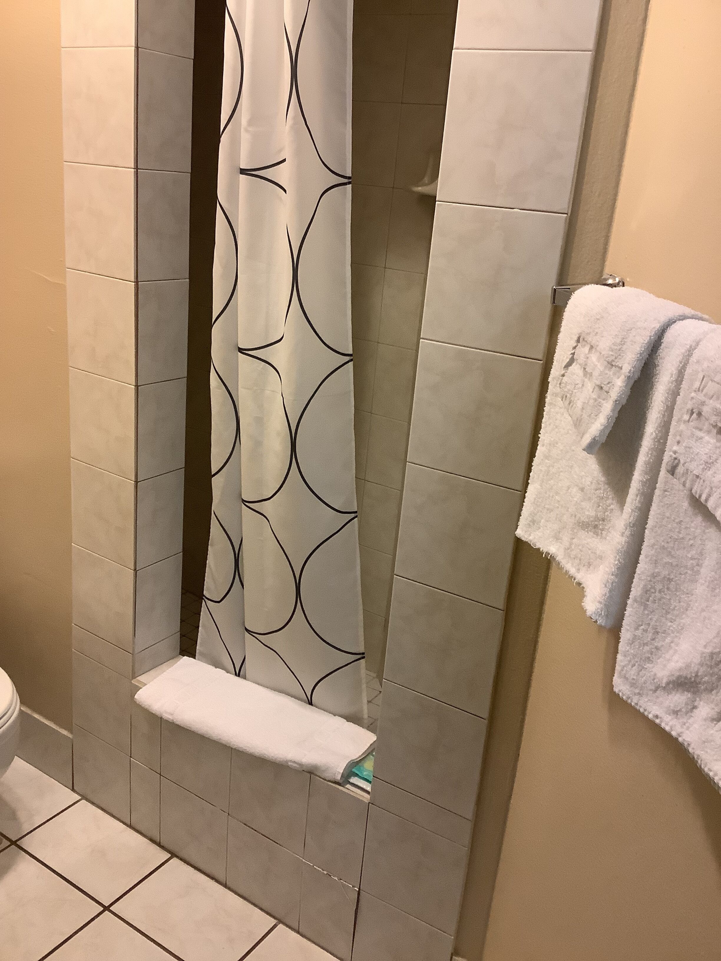 Un baño con cortina de ducha estampada con un diseño geométrico, un perchero con una toalla blanca, y paredes y suelo con azulejos.