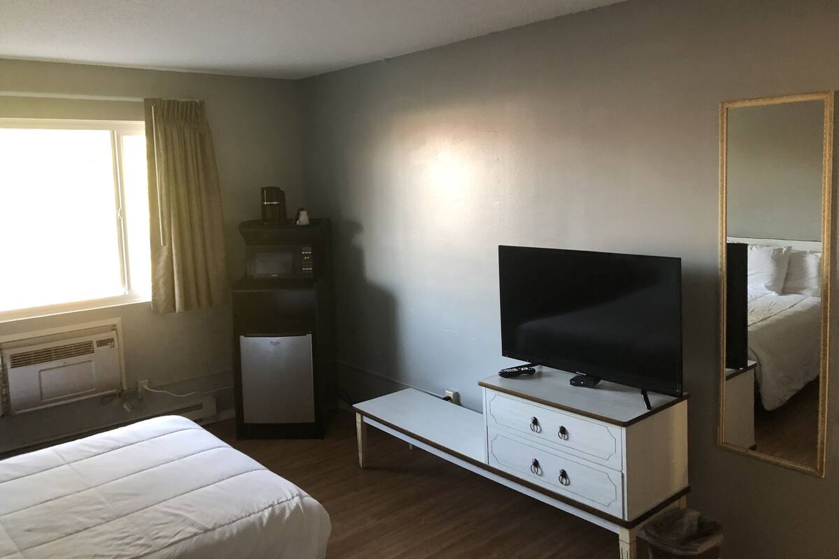A hotel room with a bed, a TV on a stand, a small table, a mirror, and a window with curtains.