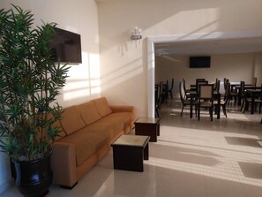 A living room with a sofa, a coffee table, and a potted plant. There is a dining area with chairs and tables in the background. - Hotel Al Akhawayn (Oujda)