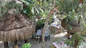 An outdoor dining area with thatched umbrellas, wooden tables, and chairs surrounded by lush greenery.