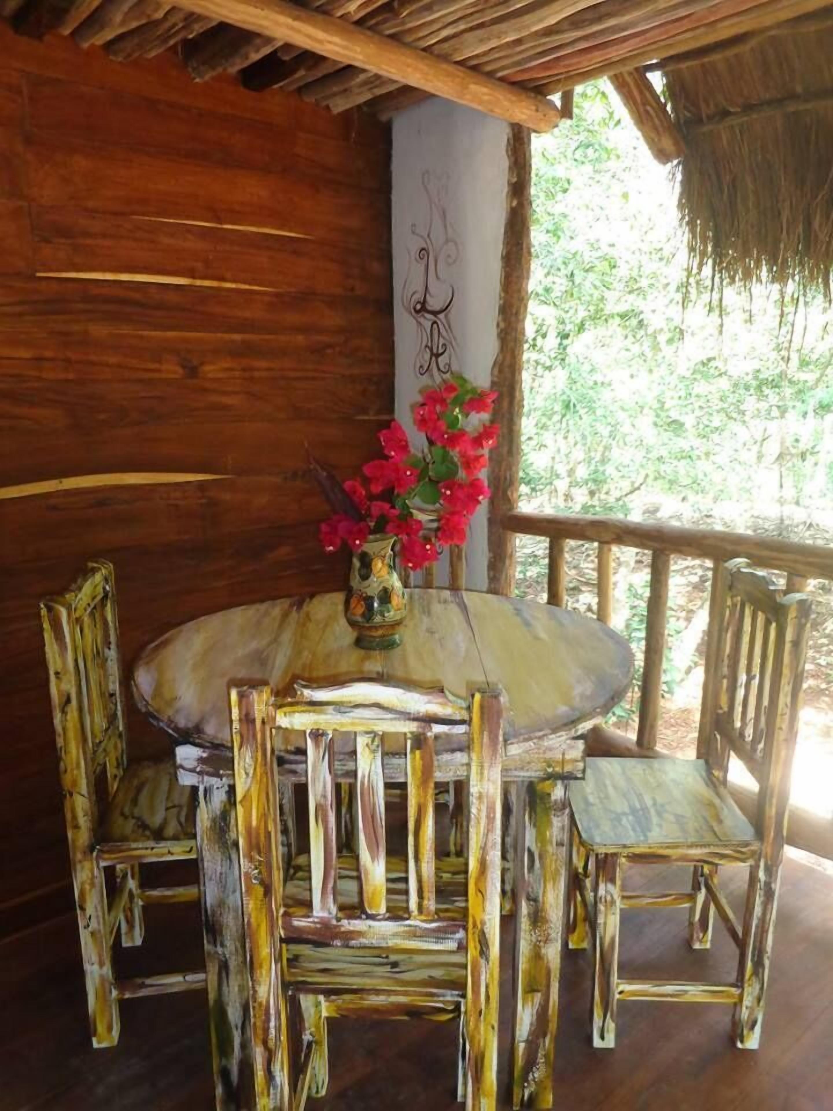 A rustic wooden table with a vase of red flowers, surrounded by wooden chairs, in a room with a thatched roof and wooden walls.