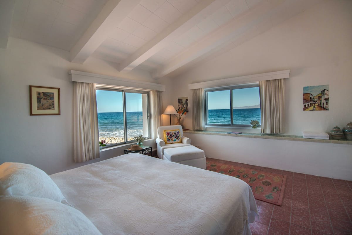 A bedroom with a large bed, two windows with ocean views, a chair, and a small table with a lamp.